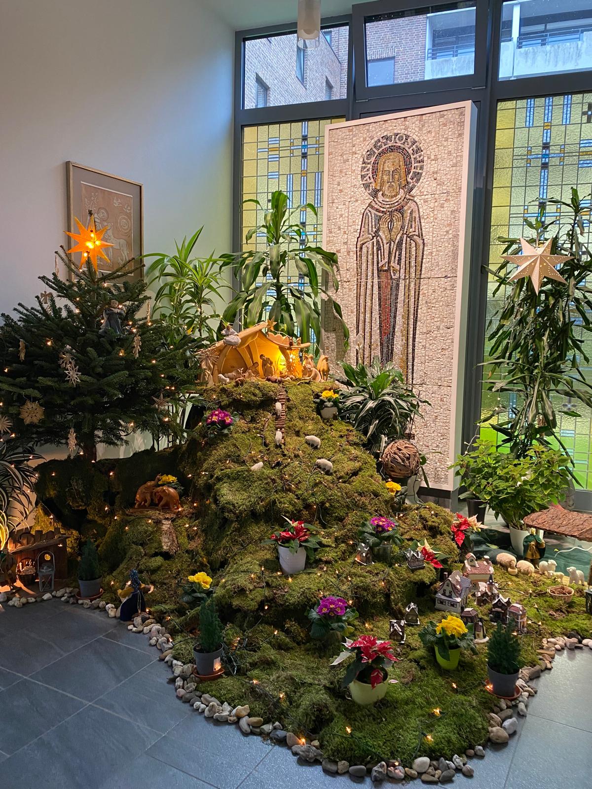Eine Krippenlandschaft mit Moos, Lichtern, Figuren, Pflanzen und Dekorationen ist auf einem Hügel in einem hellen Raum mit Kirchenfenstern und Weihnachtsbaum aufgebaut.