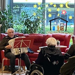 Ein älterer Mann spielt Akkordeon vor einem Publikum von Senioren, viele davon in Rollstühlen. Im Hintergrund sind ein rotes Sofa und ein Fenster mit weihnachtlicher Fensterdekoration sichtbar.
