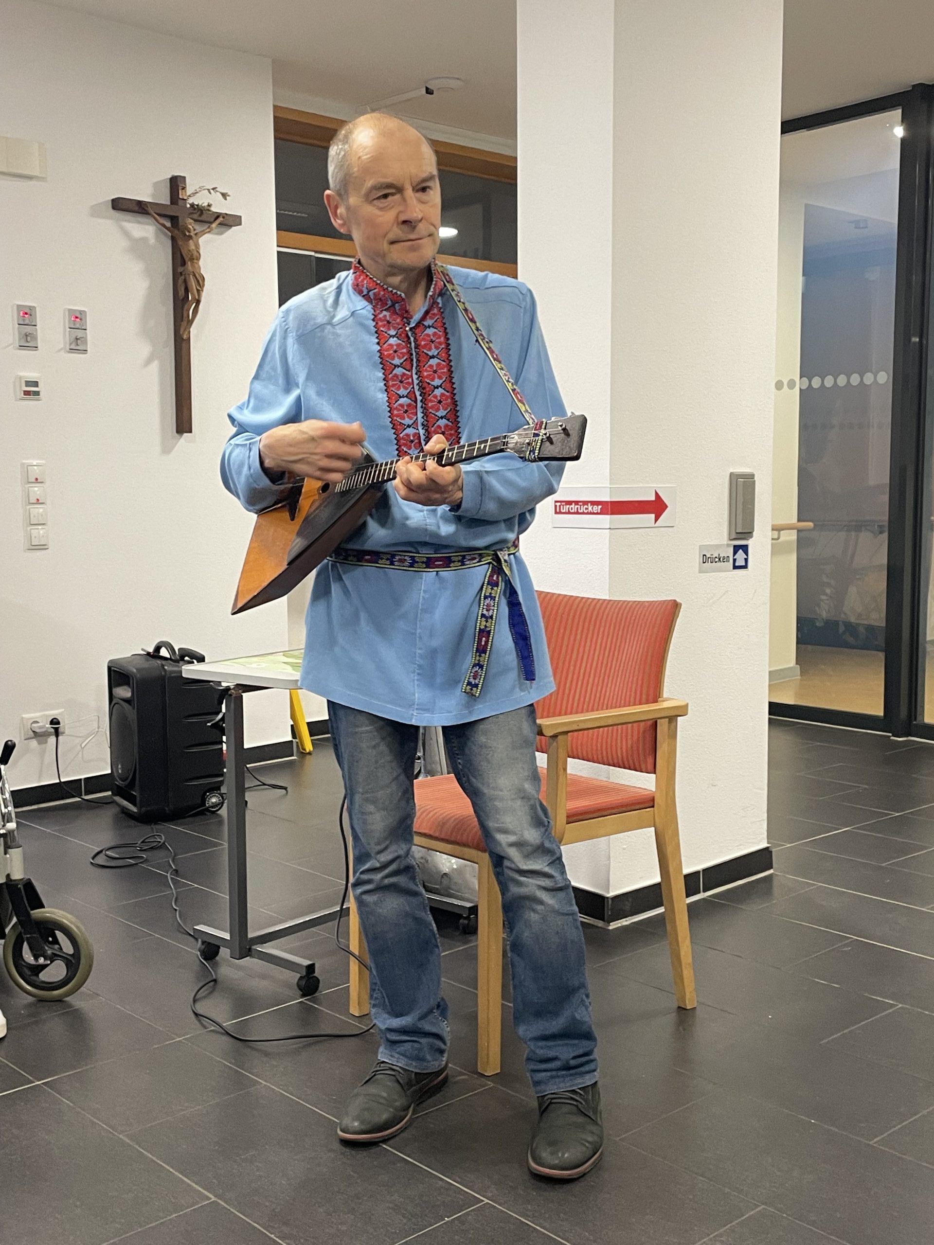 Mann spielt Balalaika im Regina Pacis Haus Ein Mann in traditioneller, blau bestickter Kleidung spielt eine Balalaika in einem Innenraum, der wie eine Pflegeeinrichtung aussieht. Hinter ihm sind ein Stuhl, ein Tisch mit Lautsprecher, ein Kreuz an der Wand und mehrere Lichtschalter zu sehen.