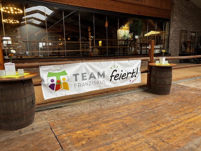 Banner „Team Franziskus feiert!“ Ein Banner mit der Aufschrift „TEAM FRANZISKUS feiert!“ hängt an einem Holzgeländer vor einer Veranstaltungslocation mit rustikalem Ambiente. Links und rechts stehen Stehtische auf alten Weinfässern mit Dekoration und Speisekarten. Im Hintergrund ist ein großer Raum mit Holzeinrichtung sichtbar.