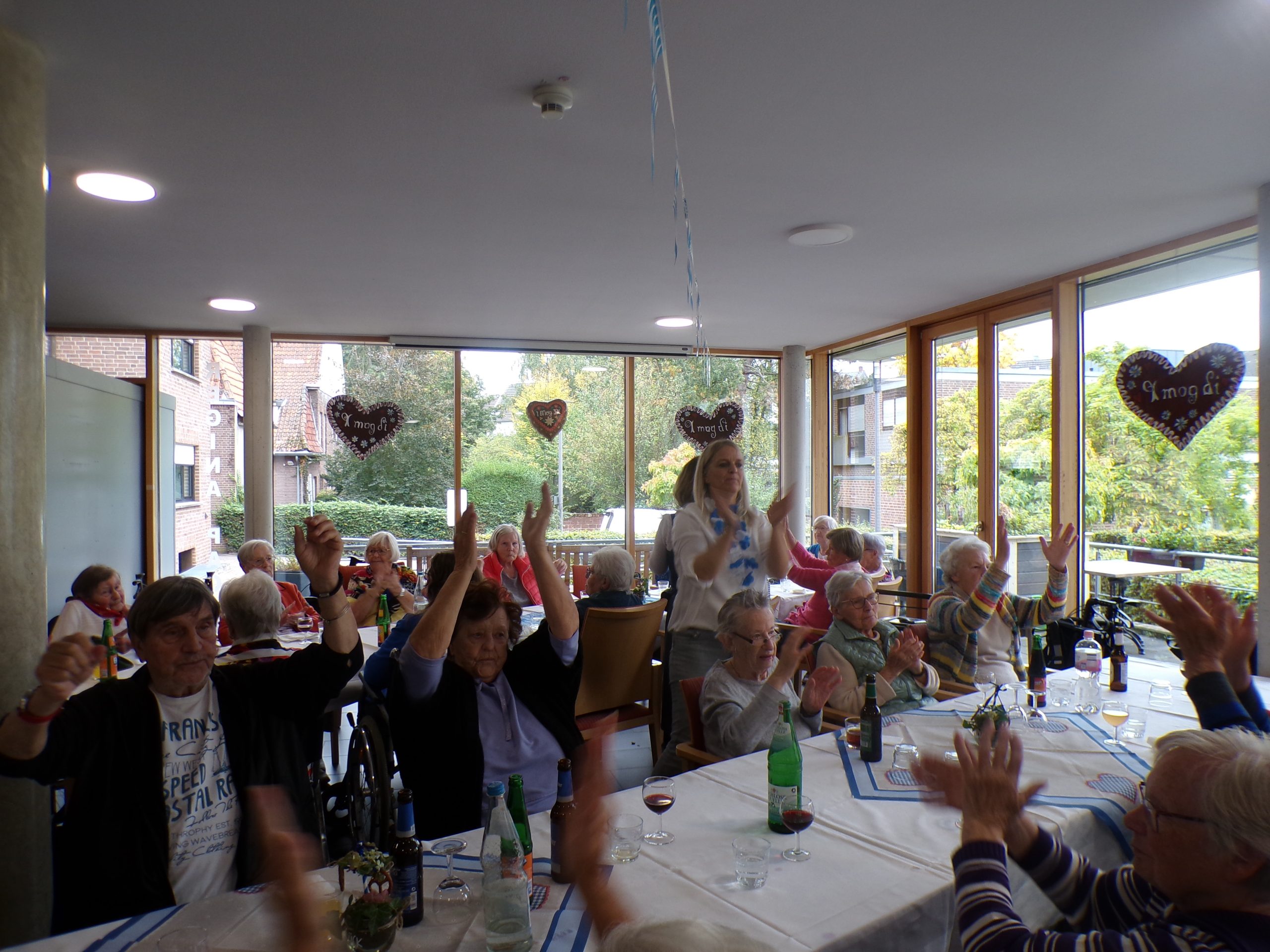 Mehrere ältere Menschen sitzen an festlich gedeckten Tischen in einem Gemeinschaftsraum und klatschen gemeinsam in die Hände. Eine jüngere Frau steht und animiert die Gruppe. Die Fenster sind mit Lebkuchenherzen dekoriert, auf denen „I mog di“ steht. Auf den Tischen stehen Getränke wie Wasser, Bier und Wein.
