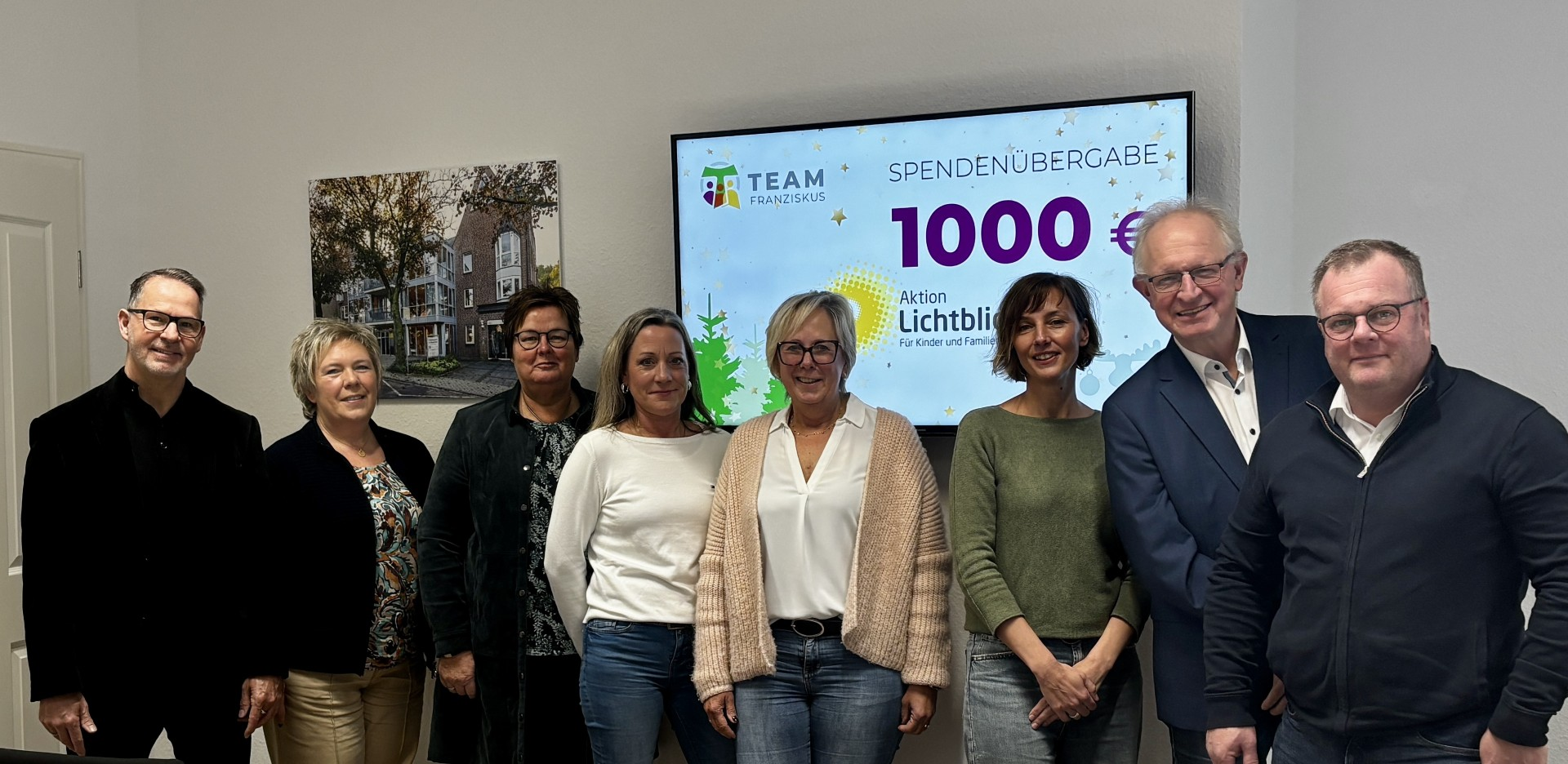 Acht Personen stehen nebeneinander in einem Raum vor einem Bildschirm, auf dem die Übergabe einer Spende von 1000 € an die „Aktion Lichtblicke“ durch Team Franziskus angezeigt wird. Alle Personen schauen in die Kamera und lächeln.