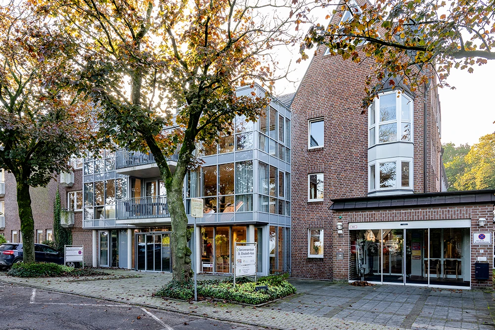 Das Bild zeigt ein modernes Seniorenzentrum mit einer Kombination aus roter Backsteinfassade und großflächigen Glasfronten. Vor dem Gebäude stehen mehrere Laubbäume mit herbstlich verfärbtem Blattwerk, ein gepflasterter Gehweg und ein Parkplatz sind ebenfalls zu sehen.