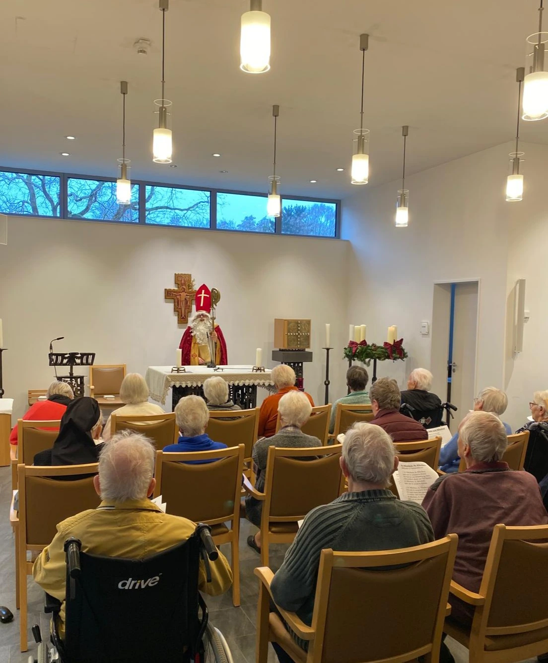 Ein als Bischof Nikolaus verkleideter Mann steht vor einem Altar in einer kleinen Kapelle und spricht zu einer Gruppe älterer Menschen, die auf Stühlen sitzen. Einige Personen sitzen im Rollstuhl, eine Ordensschwester ist ebenfalls anwesend. Der Raum ist hell beleuchtet, im Hintergrund sind ein Kreuz, Kerzen und ein Tabernakel zu sehen.
