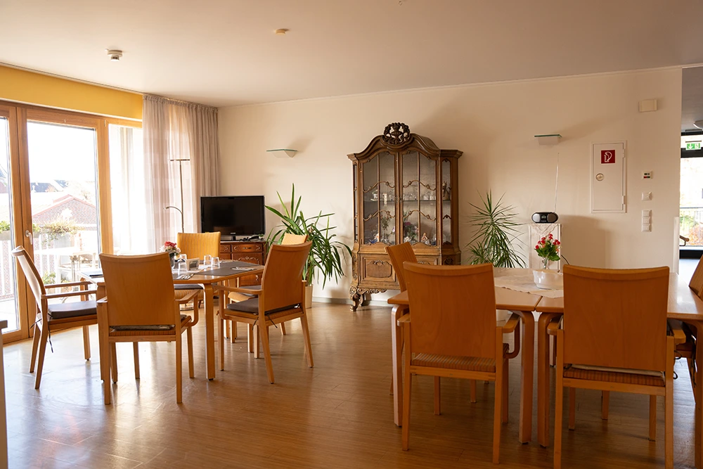 Der Innenraum zeigt einen freundlich eingerichteten Gemeinschaftsraum mit mehreren Holztischen und -stühlen, großen Fenstern mit lichtdurchlässigen Vorhängen, einem Fernseher, einem antiken Vitrinenschrank und Zimmerpflanzen. Auf den Tischen stehen kleine Blumenvasen und Trinkgläser.