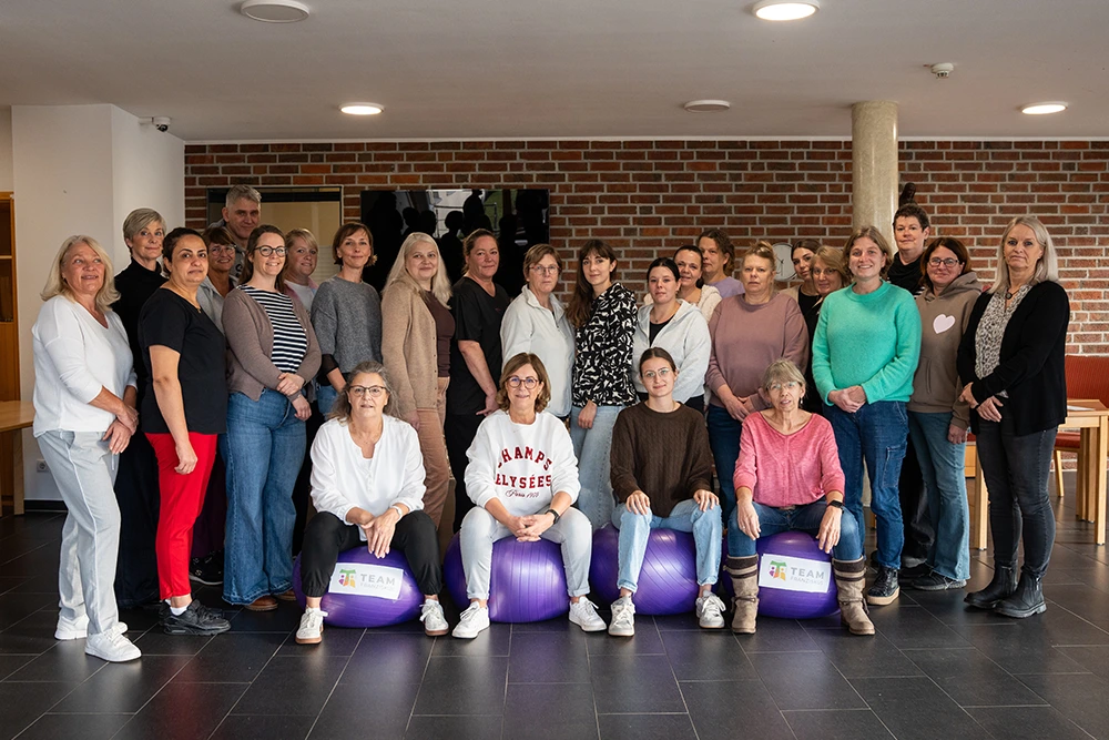 Das Bild zeigt eine größere Gruppe von Frauen und einem Mann, die als Team in einem Seniorenzentrum arbeiten. Sie stehen und sitzen in einem Innenraum mit Backsteinwand, einige sitzen auf lila Gymnastikbällen mit dem Aufdruck „Team“. Die Personen schauen in die Kamera und tragen überwiegend Freizeit- oder Arbeitskleidung.