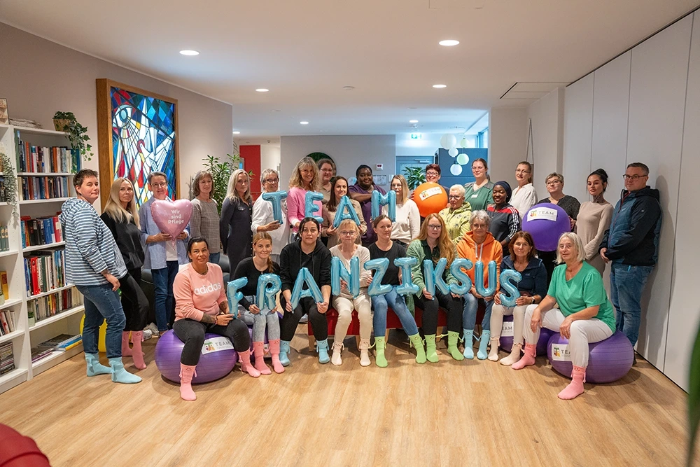 Gruppenfoto von etwa 25 Personen in einem Innenraum. Die Gruppe besteht aus Frauen und Männern verschiedener Altersgruppen. Einige sitzen auf Gymnastikbällen, andere stehen dahinter. Vorne halten Personen blaue Luftballon-Buchstaben mit der Aufschrift „TEAM FRANZISKUS“. Im Hintergrund sind Bücherregale, ein buntes Kirchenfenster und Pflanzen zu sehen.