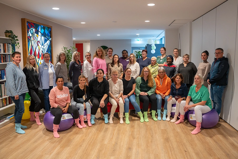 Gruppenfoto von etwa 30 Personen in einem freundlich eingerichteten Raum mit Holzfußboden. Einige sitzen auf Gymnastikbällen mit „Team Franziskus“-Logo, andere stehen dahinter. Viele tragen bunte Socken oder bequeme Kleidung. Im Hintergrund sind ein Bücherregal, ein buntes Kirchenfenster und Zimmerpflanzen zu sehen.