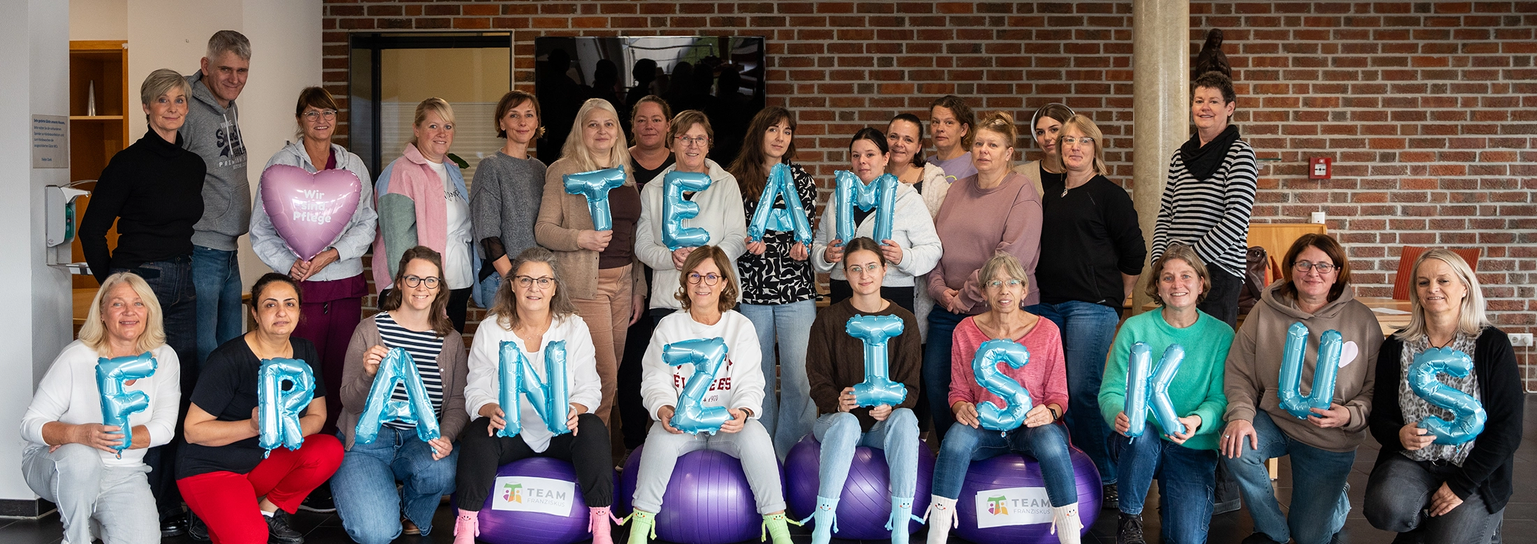 Gruppenfoto von etwa 25 Personen, die in zwei Reihen vor einer Backsteinwand sitzen und stehen. Die vordere Reihe hält blaue Ballons mit Buchstaben, die zusammen „Team Franziskus“ ergeben. Zwei Personen halten zusätzlich herzförmige Ballons mit der Aufschrift „Wir sind Pflege“. Manche sitzen auf lila Gymnastikbällen mit dem Logo „Team Franziskus“.