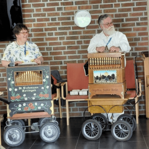 Zwei Drehorgelspieler stehen in einer Einrichtung vor einer Backsteinwand und präsentieren ihre kunstvoll gestalteten Drehorgeln auf kleinen Wagen während eines musikalischen Auftritts.