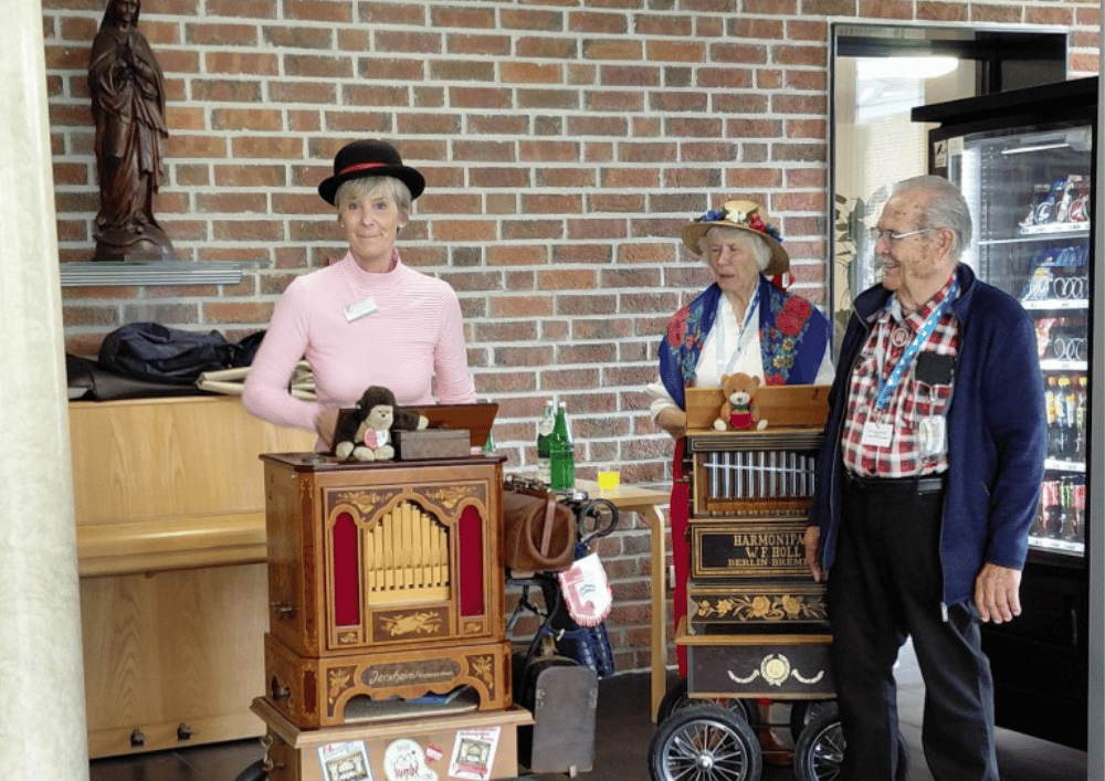 Drei Drehorgelspieler stehen in einer Einrichtung vor einer Backsteinwand und spielen auf dekorativen Drehorgeln während eines Besuchs im Rahmen der Drehorgelwallfahrt.