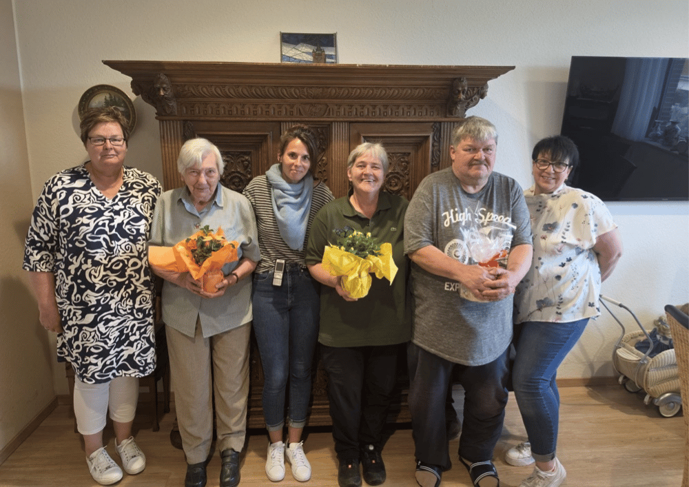 Neuer Bewohnerbeirat: Gruppenfoto mit Gratulation und Blumen Sechs Personen stehen in einer Einrichtung vor einem großen dekorativen Holzschrank, einige halten Blumensträuße in den Händen, anlässlich der Wahl des Bewohnerbeirats.