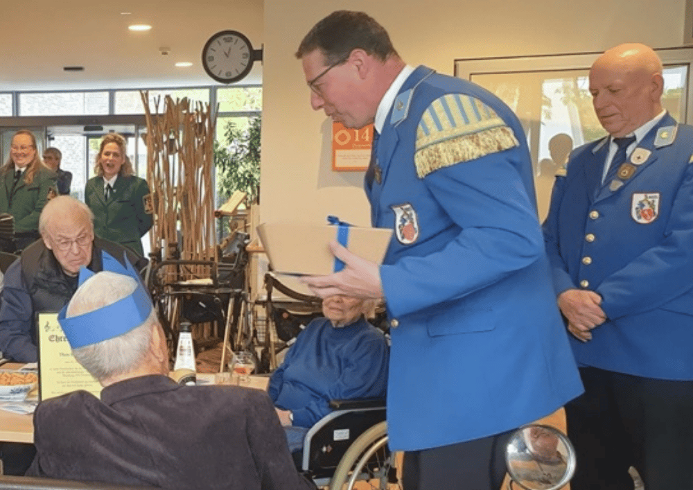 Mehrere Personen in blauen Uniformen überreichen während eines Vatertagsfrühschoppens in einer Einrichtung ein Präsent an einen Bewohner am Tisch, während weitere Anwesende zuschauen.