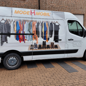 Weißer Transporter mit der Aufschrift „Modemobil“ steht vor einem Gebäude; auf der Fahrzeugseite sind bedruckte Kleiderstangen mit bunter Kleidung und Schuhen zu sehen, die das mobile Modeangebot darstellen.
