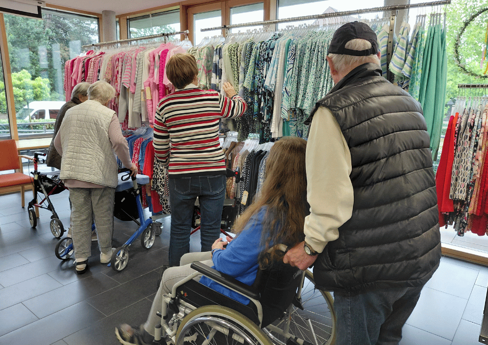 Mehrere Bewohner im Regina Pacis Haus Kevelaer schauen sich Kleidung auf mobilen Kleiderständern an; eine Person sitzt im Rollstuhl, eine andere nutzt einen Rollator, während ein Raum mit großen Fenstern als temporäre Boutique dient.