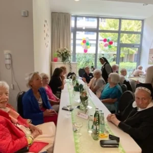 Bewohnerinnen, Mitarbeitende und eine Ordensschwester sitzen gemeinsam an einer langen, dekorierten Festtafel.