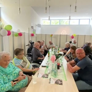 Bewohnerinnen, Bewohner und Mitarbeitende sitzen gemeinsam an einer langen Tafel in einem mit Luftballons geschmückten Raum.