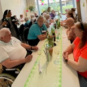 Bewohnerinnen, Bewohner und Mitarbeitende sitzen gemeinsam an einer langen, festlich dekorierten Tafel und unterhalten sich.