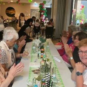 Bewohnerinnen, Bewohner und Mitarbeitende sitzen an einer langen, festlich dekorierten Tafel und applaudieren.