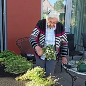 Eine ältere Frau schneidet an einem Gartentisch verschiedene frische Kräuter für die weitere Verarbeitung zurecht.