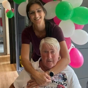 Eine Mitarbeiterin umarmt einen sitzenden Bewohner vor einer Dekoration aus bunten Luftballons.