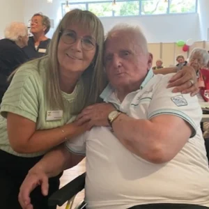 Eine Mitarbeiterin legt den Arm um einen sitzenden Bewohner und posiert mit ihm für ein gemeinsames Foto.