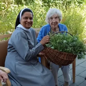 Zwei Frauen sitzen auf einer Terrasse im Grünen und halten gemeinsam einen großen Korb mit frisch gesammelten Kräutern.