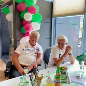 Zwei Bewohner sitzen gut gelaunt an einer festlich gedeckten Tafel vor einer Dekoration aus bunten Luftballons.
