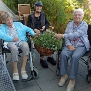Drei ältere Frauen sitzen im Garten um einen Korb mit frisch gesammelten Kräutern.