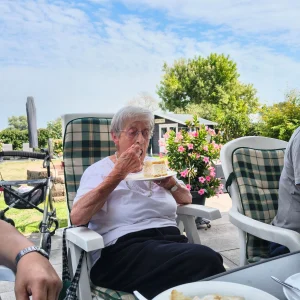 Eine ältere Frau sitzt im Garten und isst ein Stück Torte. Im Vordergrund steht ein weiterer Teller mit Kuchen.