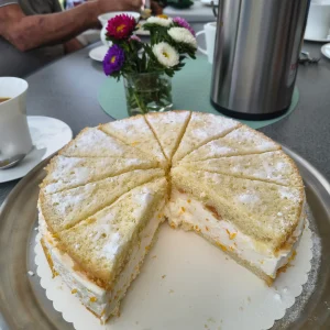 Eine angeschnittene Sahnetorte steht auf einer Kaffeetafel, daneben ein kleiner Blumenstrauß und eine Thermoskanne.