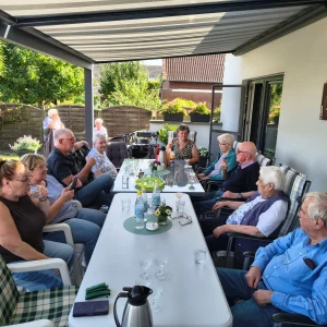 Mehrere ältere Menschen und Begleitpersonen sitzen gemeinsam an einer langen Kaffeetafel auf einer überdachten Terrasse.