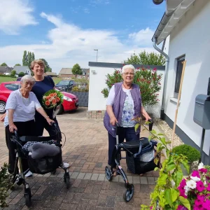 Zwei ältere Frauen mit Rollatoren und eine Begleitperson stehen vor einem Wohnhaus. Die Begleitperson hält einen Blumenstrauß.