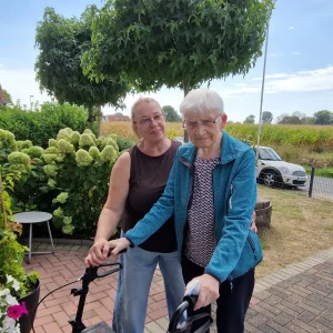 Eine ältere Frau steht mit einem Rollator im Garten. Eine weitere Frau begleitet sie und steht unterstützend hinter ihr.