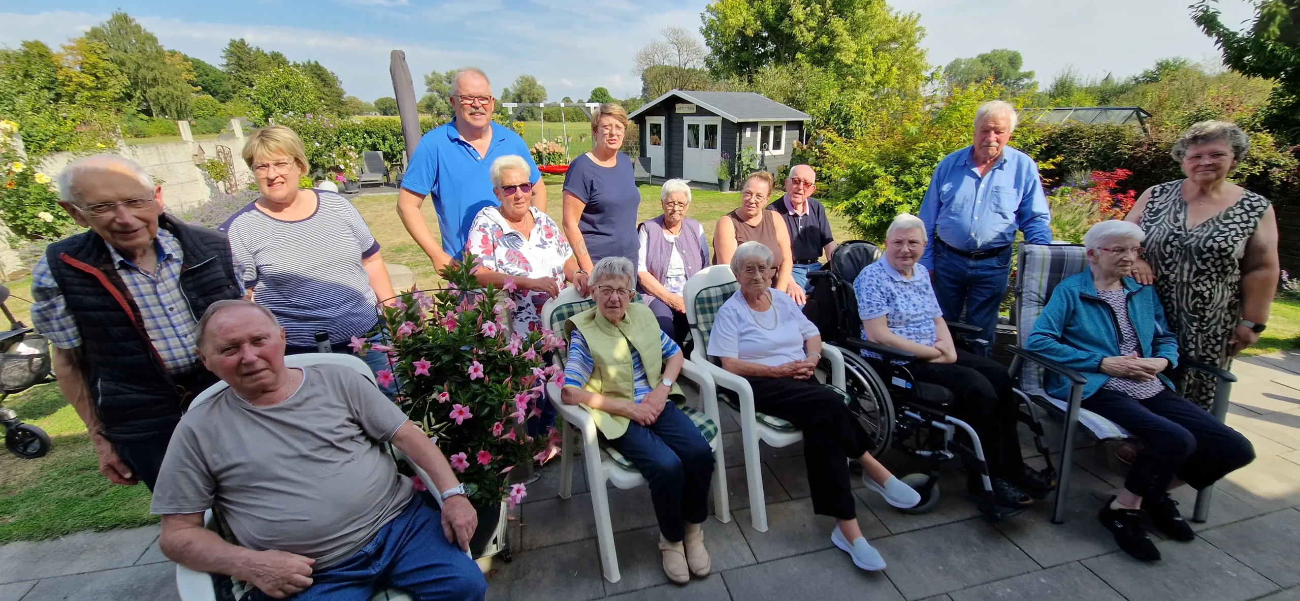 1000087442 Eine Gruppe älterer Menschen und Begleitpersonen versammelt sich bei sonnigem Wetter für ein gemeinsames Foto im Garten.“
