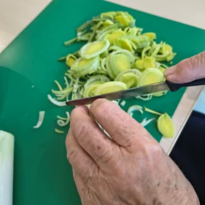 Nahaufnahme von Händen, die Lauch mit einem Messer auf einem grünen Schneidebrett in Ringe schneiden.