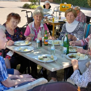 Bewohnerinnen und Bewohner sitzen im Schatten zusammen und genießen das gemeinsame Essen beim Sommerfest.