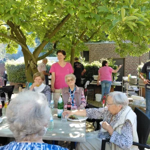 Bewohnerinnen, Bewohner und Mitarbeitende sitzen gemeinsam unter einem Sonnenschirm beim Essen.