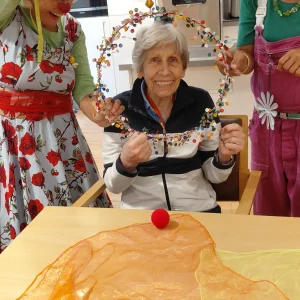 Eine ältere Frau lächelt in die Kamera und hält gemeinsam mit zwei Clowninnen einen mit bunten Knöpfen geschmückten Reifen.