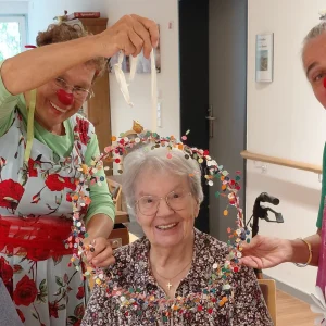 Eine ältere Frau sitzt zwischen zwei Clowninnen mit roten Nasen und lächelt durch einen bunt geschmückten Reifen