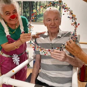 Ein älterer Mann sitzt in einem Stuhl, während eine Clownin mit roter Nase einen bunt geschmückten Reifen vor ihn hält.