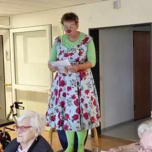 Eine als Clown verkleidete Frau steht auf einem Stuhl und liest etwas vor.