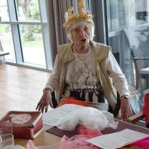Eine ältere Frau mit einer Plastikkrone auf dem Kopf sitzt auf einem Stuhl.