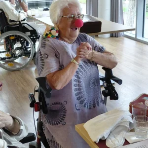 Eine ältere Frau im Rollstuhl trägt eine rote Clownsnase und lächelt mit gefalteten Händen.