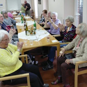 Ältere Menschen sitzen an einer langen, herbstlich dekorierten Tafel und klatschen. Im Hintergrund tanzt ein Paar.