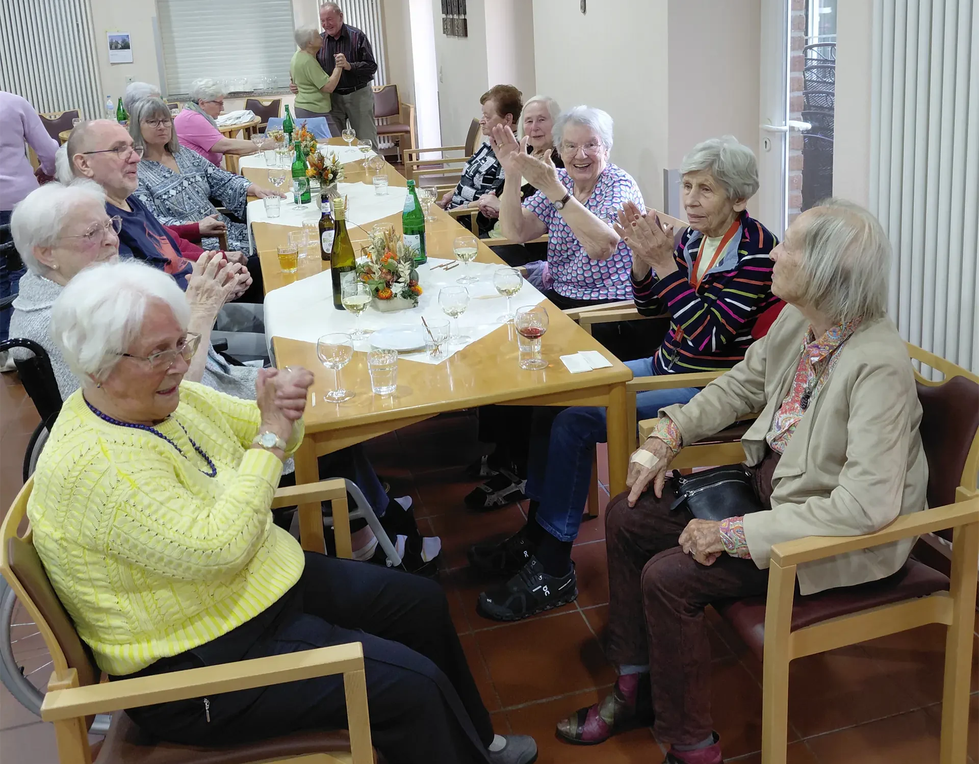 Geselliges Beisammensein mit Tanz und Applaus Ältere Menschen sitzen an einer langen, herbstlich dekorierten Tafel und klatschen. Im Hintergrund tanzt ein Paar.