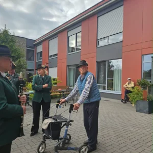 Ein älterer Mann mit Hut und Rollator steht neben Mitgliedern einer Blaskapelle im Innenhof.