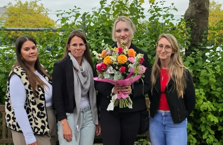 67e34c5b-7b66-48e4-9c17-d70f30d2ad83 Vier Frauen stehen gemeinsam im Freien vor einer grünen Hecke. Die Frau in der Mitte hält einen großen, bunten Blumenstrauß.