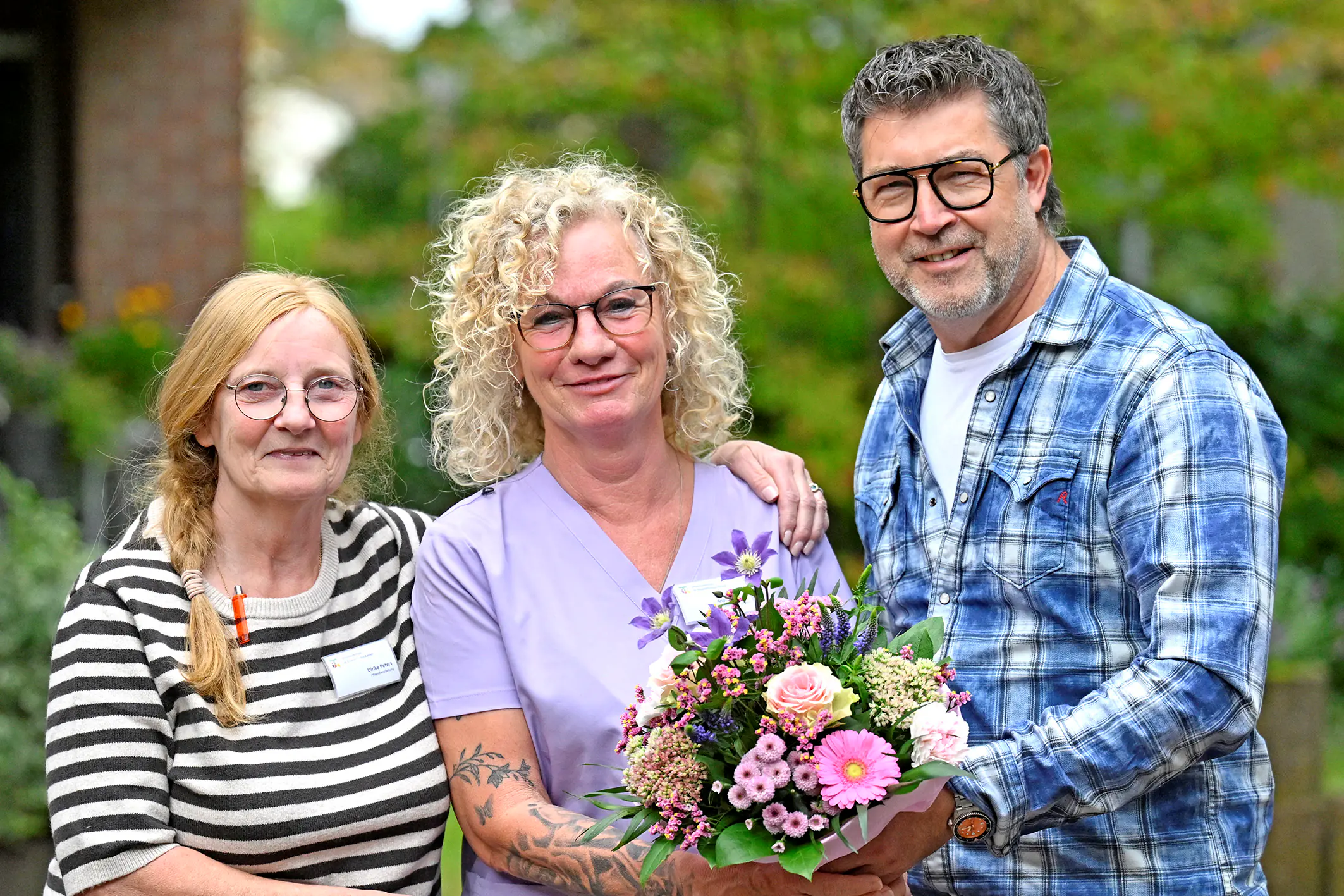 Andrea Rosner feiert ihren Abschluss in der Pflegefachassistenz Eine Frau in lila Pflegekleidung hält einen Blumenstrauß. Neben ihr stehen eine Frau und ein Mann. Alle drei lächeln in die Kamera.