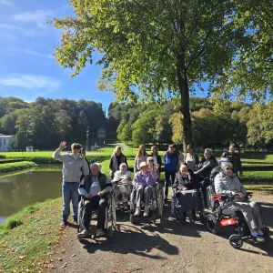 Eine Ausflugsgruppe mit älteren Menschen in Rollstühlen und Begleitpersonen posiert an einem Teich. Einige winken in die Kamera.